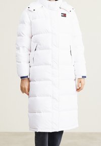 Vit vadderad lång kappa med hög krage och dragkedjestängning. Har dubbla sidofickor och en logotyp patch på bröstet. Slät textur.