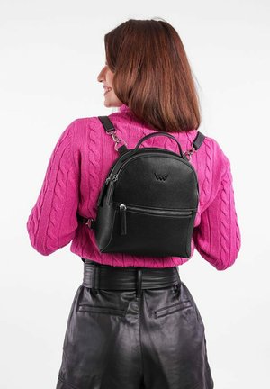 Černý kožený mini batoh ve kulatém tvaru, s přední kapsou na zip a nastavitelnými ramenními popruhy, nošený přes růžový svetr s pleteným vzorem.