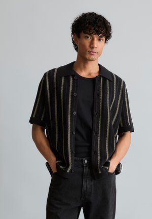 Junger Mann mit lockigem Haar, der ein kurzärmeliges Hemd mit schwarzen vertikalen Streifen über einem schwarzen T-Shirt trägt, Hände in den Taschen, neutraler Hintergrund.