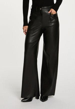 Donna che indossa pantaloni in pelle neri a gamba larga con vita alta e tasche frontali, abbinati a stivali neri con punta a stiletto e un top nero a coste.