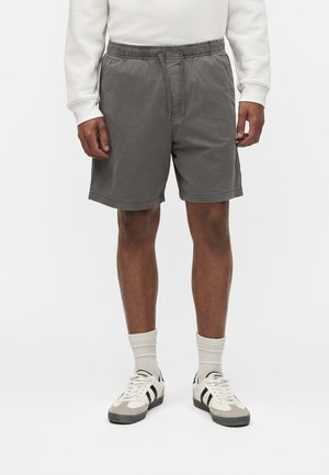 Mężczyzna ubrany w szare spodenki z paskiem, białe skarpetki za kostkę, białe i czarne sneakersy oraz biały top z długim rękawem, stojący na jednolitym tle.