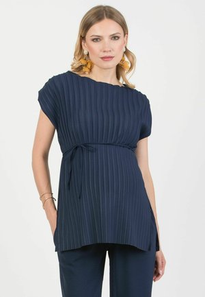 Femme portant une tunique plissée bleu marine avec un lien sur le côté, pantalon assorti et grandes boucles d'oreilles florales jaunes, debout sur fond blanc.