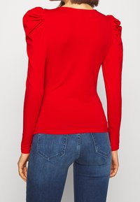 Maglietta rossa a maniche lunghe con spalle a palloncino, design aderente e texture liscia. Indossata con jeans blu, dotati di tasche posteriori e fit a vita media.