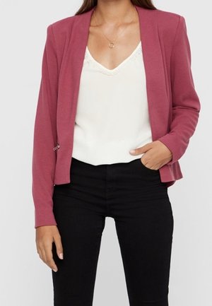 Femme portant un blazer rose, un caraco blanc orné de dentelle et un pantalon noir, une main dans la poche et un collier doré.