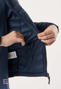 Mains tenant ouvert l'intérieur d'une veste matelassée bleu marine, révélant une poche intérieure et une étiquette d'entretien.