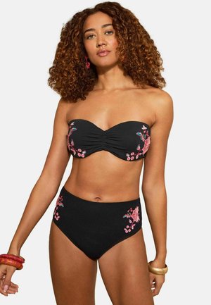 Kvinde med krøllet hår iført sort, stropløs bikini med pink blomstermønster og kraftige armbånd på begge håndled.