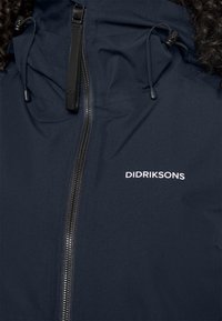 Veste imperméable marine avec fermeture éclair à l'avant, capuche à cordon et détails de logo. Texture de tissu lisse ; comporte des zips et des poignées noires.