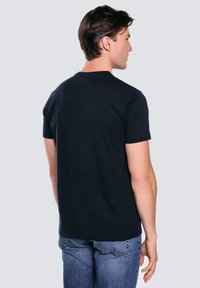 Mężczyzna o ciemnych włosach, ubraną w prosty czarny t-shirt z krótkim rękawem i niebieskie jeansy, stojący tyłem, aby pokazać tył ubrania.