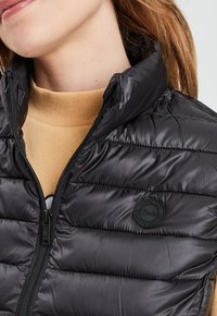 Zwarte puffer vest met ritssluiting, gestikt ontwerp, hoge kraag en een ronde logopatch. Draagt over een beige coltrui.