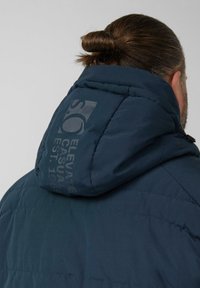 Granatowa, pikowana kurtka z dużym kapturem. Na plecach znajduje się szare logo, tkanina o fakturze z detalami przeszyć.