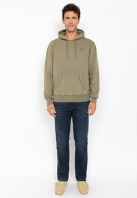 Oliwkowy hoodie z kieszenią z przodu, regulowanymi sznurkami i ściągaczami przy rękawach, w połączeniu z ciemnoniebieskimi dżinsami i beżowymi butami typu slip-on.