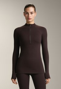 Top marrone a maniche lunghe con colletto alto e mezza zip. Presenta un tessuto testurizzato e un design aderente, adatto per l'abbigliamento sportivo.