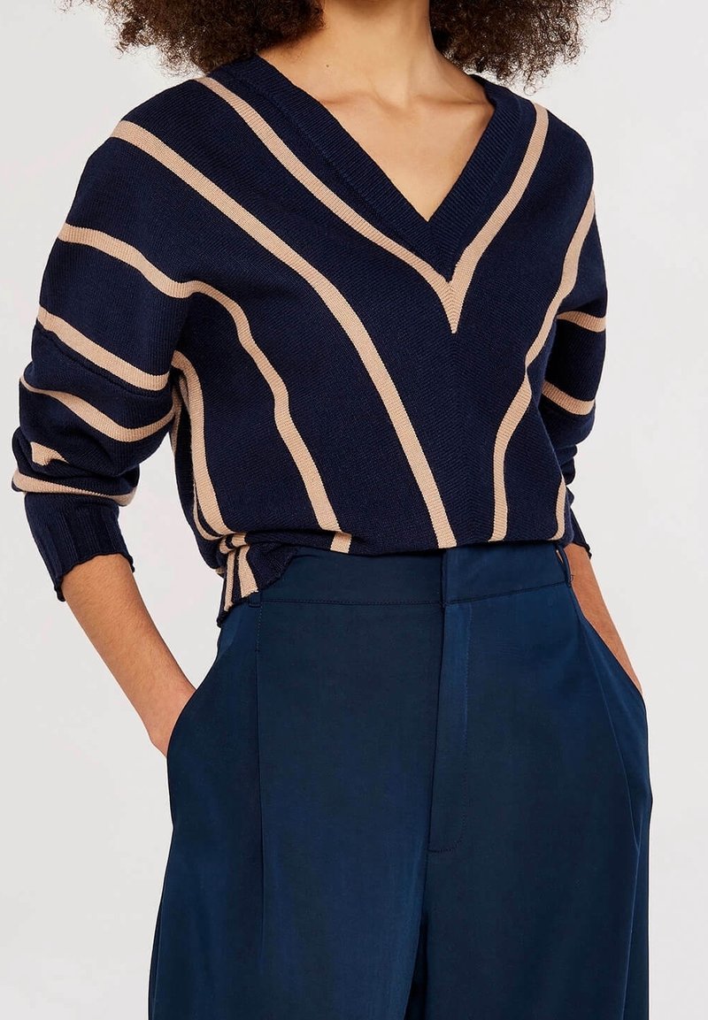 Personne portant un pull en V bleu marine avec des rayures diagonales beige et un pantalon taille haute assorti bleu marine, les mains dans les poches.