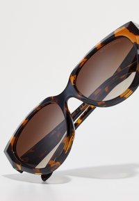 Occhiali da sole tartaruga con forma geometrica a occhio di gatto, lenti scure a gradiente e una montatura in acetato lucido. Branding dettagliato di Michael Kors all'interno.