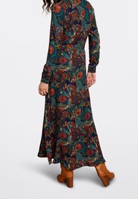 Kvinna klädd i en färgglad klänning med blommönster och abstrakt mönster, långa ärmar och tanfärgade klackstövletter, stående mot vit bakgrund.