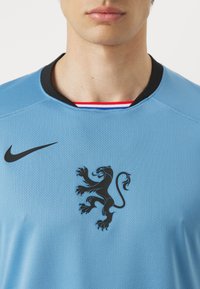 Maglietta sportiva blu chiaro con dettagli neri. Presenta un emblema di un leone nero sul petto e un colletto a girocollo nero. Tessuto testurizzato.