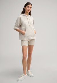 Hoodie beige clair à manches courtes, avec une poche avant et le logo "JOOP !" ; associé à un short assorti et des baskets blanches, offrant une texture douce.