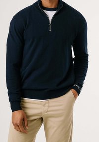 Pull en tricot bleu marine avec col montant zippé, poignets et ourlet côtelés, orné d'un logo sur le poignet, associé à un pantalon beige clair.