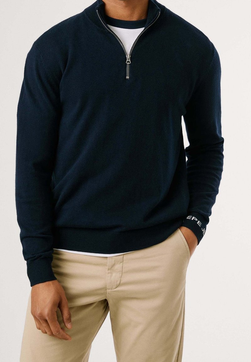 Pull en tricot bleu marine avec col montant zippé, poignets et ourlet côtelés, orné d'un logo sur le poignet, associé à un pantalon beige clair.