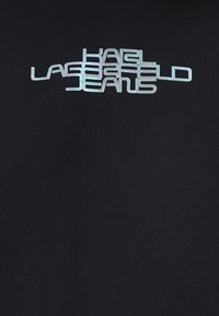 Crna majica od tkanine s reflektirajućim srebrnim logotipom koji glasi "KARL LAGERFELD JEANS" u modernom, stiliziranom fontu.