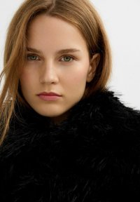 Mujer joven con cabello castaño claro, ojos verdes y maquillaje natural, vestida con un abrigo negro esponjoso frente a un fondo neutro.