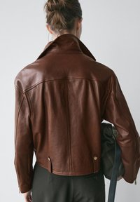 Giacca in pelle marrone con collo alto, design aderente, maniche con zip e dettagli in metallo. Indossata sopra pantaloni scuri, con una borsa in mano.