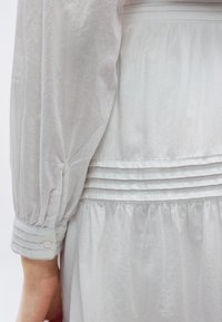 Bijeli rukav haljine s nabranim manžetom i detaljem s gumbom, uz plisirani pojas i tkaninu suknje koja slobodno pada.