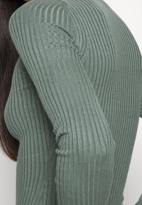 Geribbelde langemouwen top in een gedempte groene stof, met een aansluitend ontwerp en textuurdetails langs de schouders en mouwen.