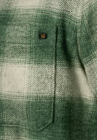 Grünes und cremefarbenes kariertes Flanellgewebe mit einer strukturierten Oberfläche. Verfügt über einen schwarzen Hardware-Akzent auf einer Brusttasche.