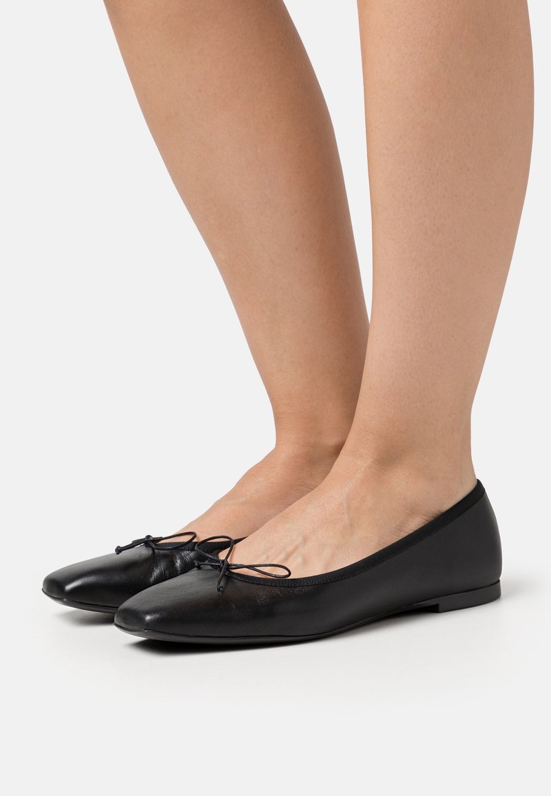 Paire de ballerines en cuir noir avec petits nœuds, portées pieds nus sur un fond clair uni.