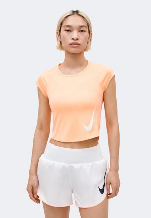 Ung person med kort blondt hår iført en ferskenfarvet cropped atletisk top og hvide Nike-shorts med sort logo på ensfarvet baggrund.