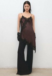 Top de satén marrón con ribete de encaje negro, dobladillo asimétrico y tirantes delgados, combinado con pantalones negros de talle alto y guantes negros.