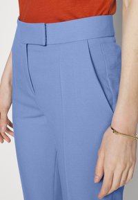 Ljusblå skräddarsydda byxor med bred midjeband, veck framtill och sidofickor, kombinerat med en röd topp och ett guldarmband på handleden.