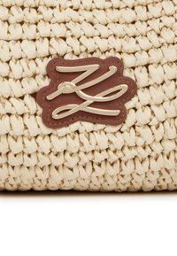 Splotkana torba ze słomy w kolorze kremowym o teksturowanej powierzchni; wyposażona w brązową skórzaną metkę z kursywnym metalowym akcentem logo.