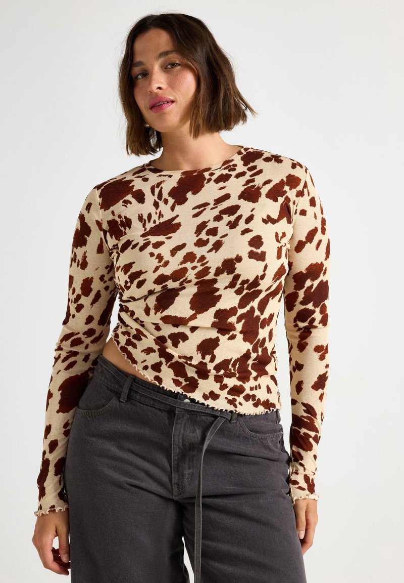 Donna che indossa un top a maniche lunghe beige e marrone maculato con pantaloni grigi scuri a vita alta su uno sfondo bianco semplice.