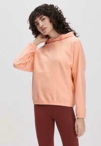 Pfirsichfarbener, knapper Hoodie aus weichem Stoff mit langen Ärmeln, ausgestattet mit einer Kordelzugkapuze und seitlichen Taschen, kombiniert mit braunen Leggings.