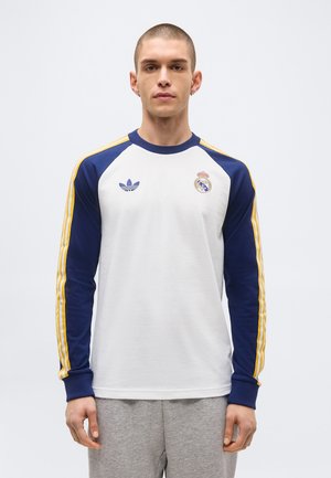 Jeune homme portant un maillot de sport à manches longues blanc et bleu marine avec des bandes jaunes, logos Adidas et Real Madrid, debout devant un fond uni.