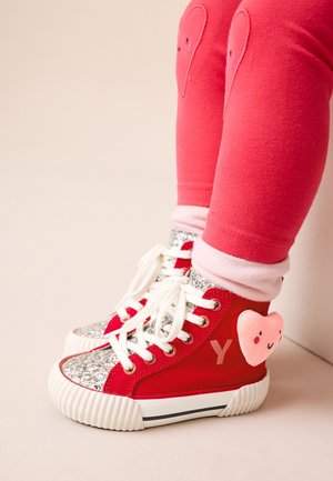 Kind trägt rote High-Top-Sneaker mit silberglitzernden Spitzen und einem lächelnden pinken Herzpatch, kombiniert mit roten Leggings mit Herzpatches.