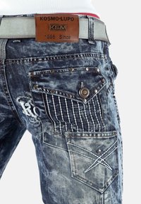 Gros plan sur un jean en denim bleu délavé avec des coutures décoratives, des clous métalliques, et une pièce de ceinture en cuir marron portant l'inscription "KOSMO LUPO K&M 1996 Since."