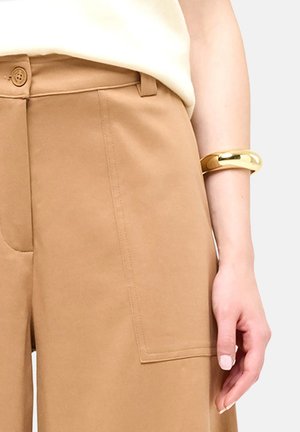 Primo piano di una persona che indossa pantaloni beige con un bottone, una maglietta bianca e un braccialetto d'oro al polso.