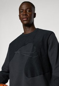 Black sweatshirt with textured patches, featuring a prominent Nike logo in light stitching. Model holds a basketball.