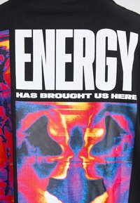 Svart bomull t-skjorte med fet hvit tekst "ENERGIEN HAR FØRT OSS HIT" over et levende, abstrakt grafikk i røde, blå og gule nyanser.