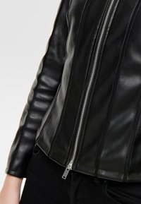 Veste en cuir noir au design ajusté, avec une fermeture éclair centrale, des coutures verticales et une texture côtelée le long des manches.