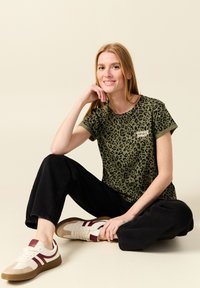 Camiseta verde oliva con estampado de leopardo negro y texto "dancing queen", combinada con pantalones negros holgados y zapatillas blancas con acentos burdeos.