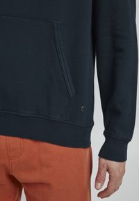 Tumši zila sporta jaka ar priekšējo kabatu, ribinātu piedurkņu un apakšmalu, kā arī mazu logotipa detaļu netālu no apakšmalas. Saskaņota ar oranžiem biksēm.