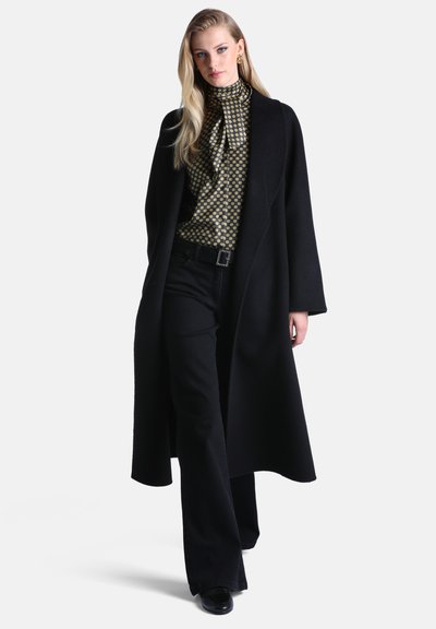 Femme aux longs cheveux blonds portant un chemisier à col haut à motif doré, un long manteau noir, un pantalon noir et des chaussures noires, marchant vers l'avant sur fond blanc.