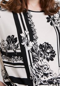 Fladdrande topp i krämvit med svart blommönster. Har djärva blommiga mönster och geometriska linjer. Slät textur med rund halsringning.