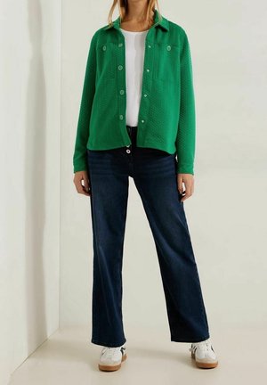 Persoon draagt een groene jas met knopen over een witte top, donkerblauwe spijkerbroek en witte sneakers, staand tegen een witte achtergrond.