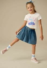 Barn som bär en vit t-shirt med en färgglad "Stitch"-grafik, tillsammans med en blå denimkjol och rosa sneakers med vita och blå randiga strumpor.