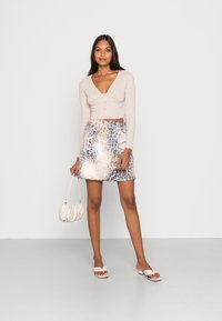 Top cropped texturizado bege com decote em V e botões, combinado com uma saia mini estampada em tons de bege e cinza, e uma pequena mala creme.